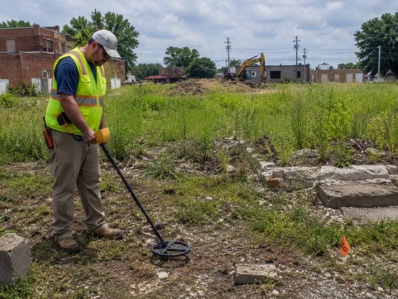 Land surveyor searching for a buried property corner marker on an overgrown vacant lot before redevelopment.